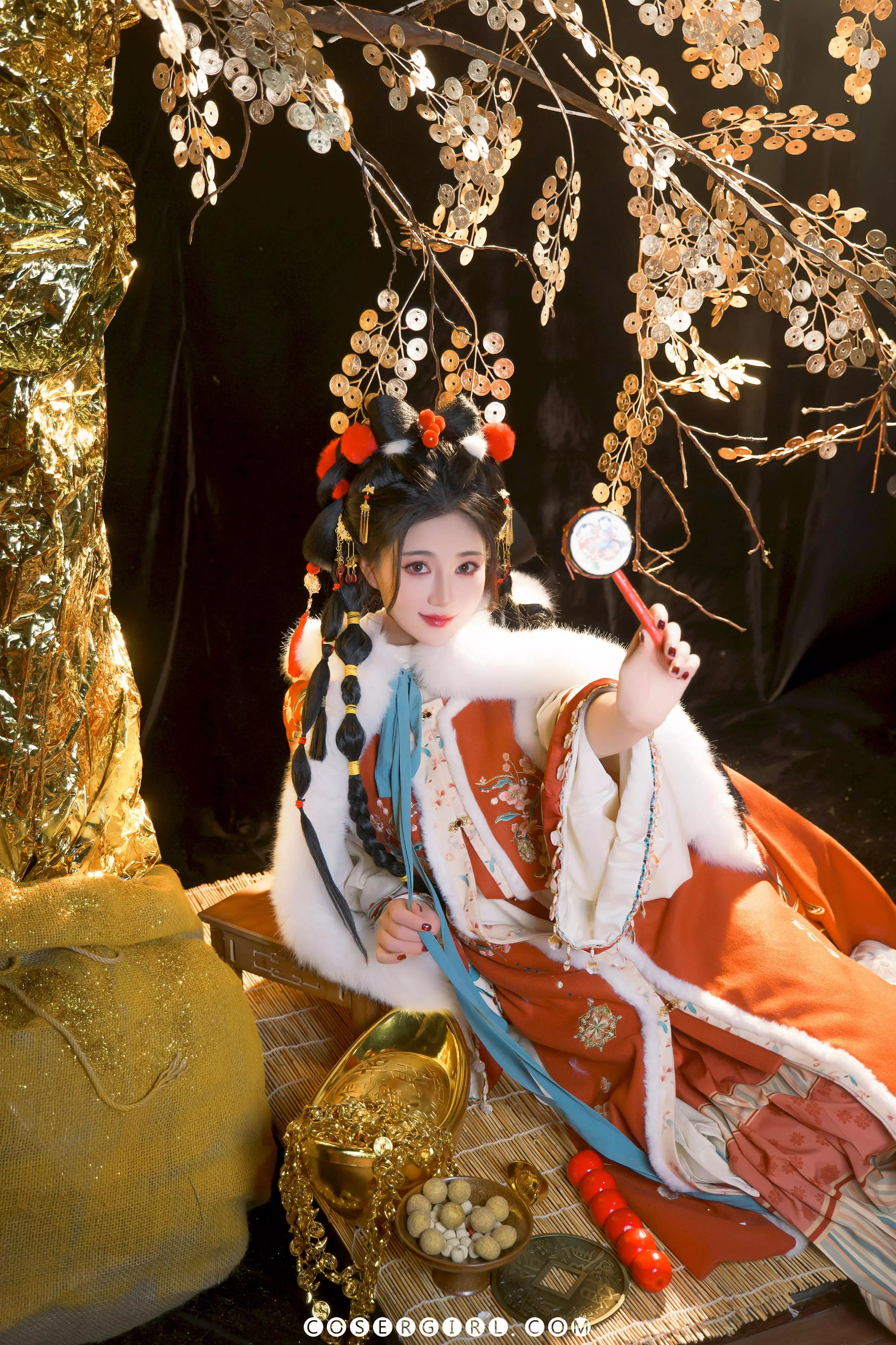 2025年宝坐在竹席上,手持一个带有铜钱图案的红色小镜子,侧头看向镜头。