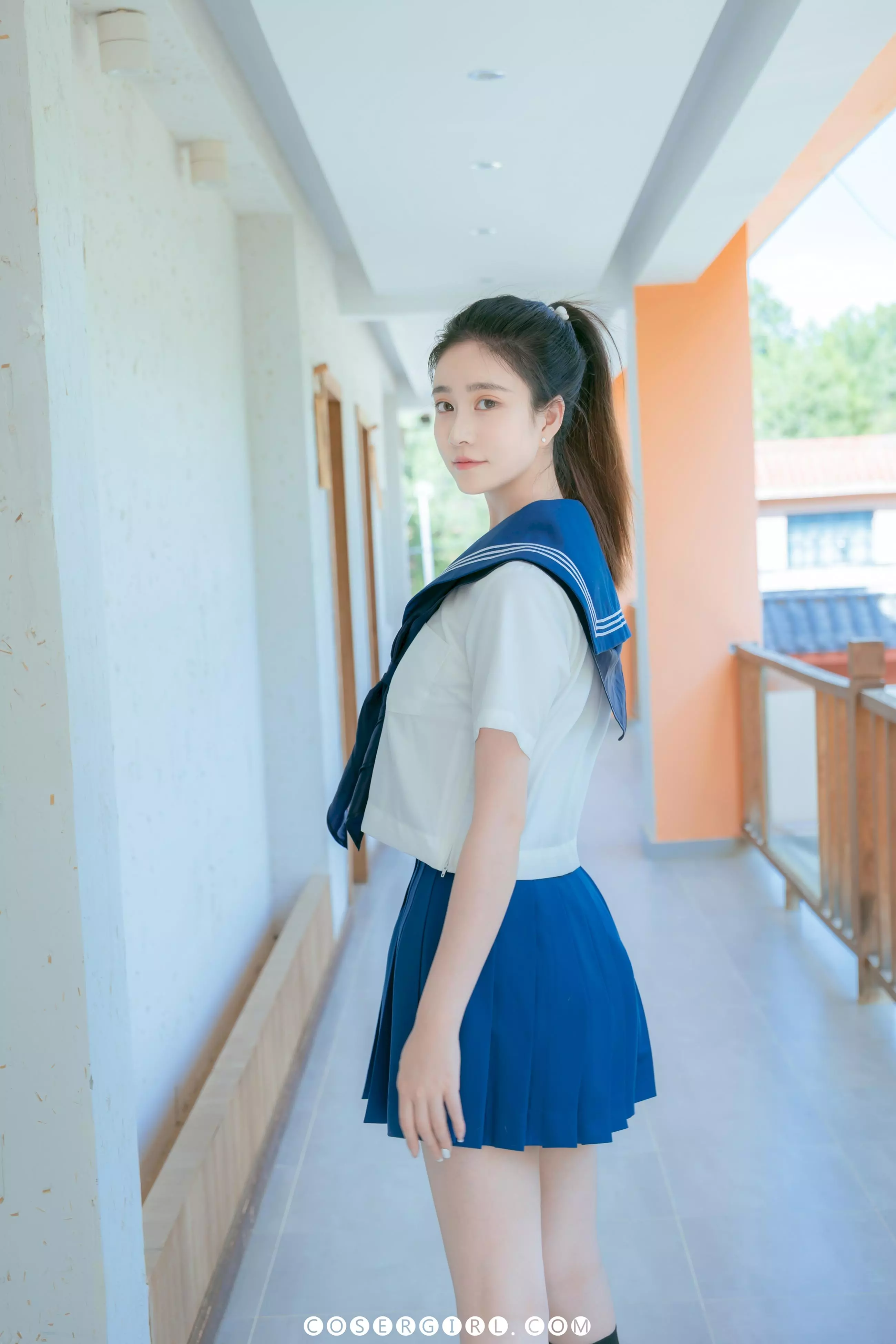 年年穿着水手服和百褶裙,站在白色走廊里,侧身回眸看着镜头。