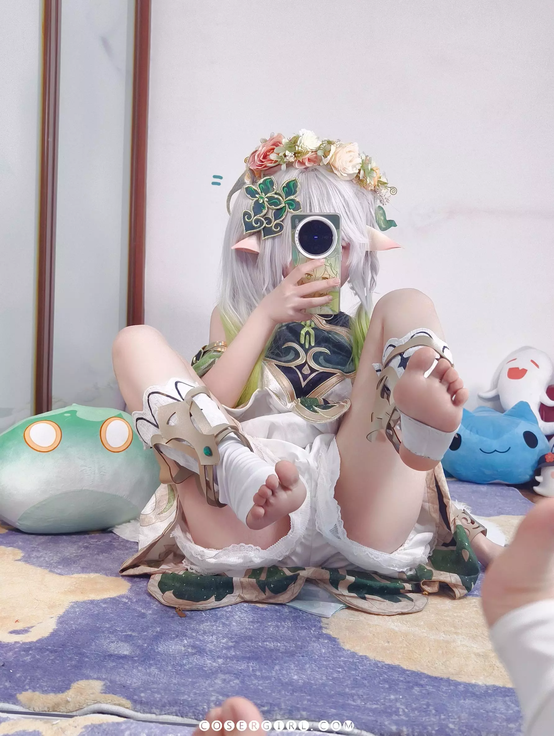 ran3扮演的纳西妲侧坐在地毯上,双手举着手机自拍,背景是她的床和柜子。