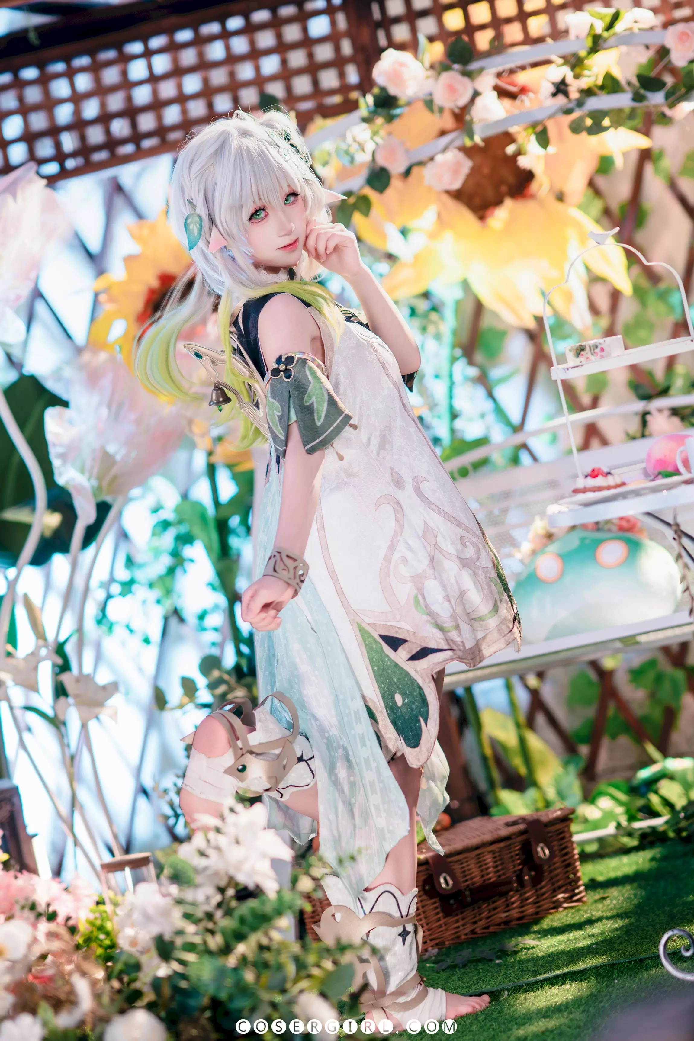 ran3扮演的纳西妲单脚站立在花园场景中,一手轻抚脸颊,另一只手拿着飘带,显得活泼又俏皮。