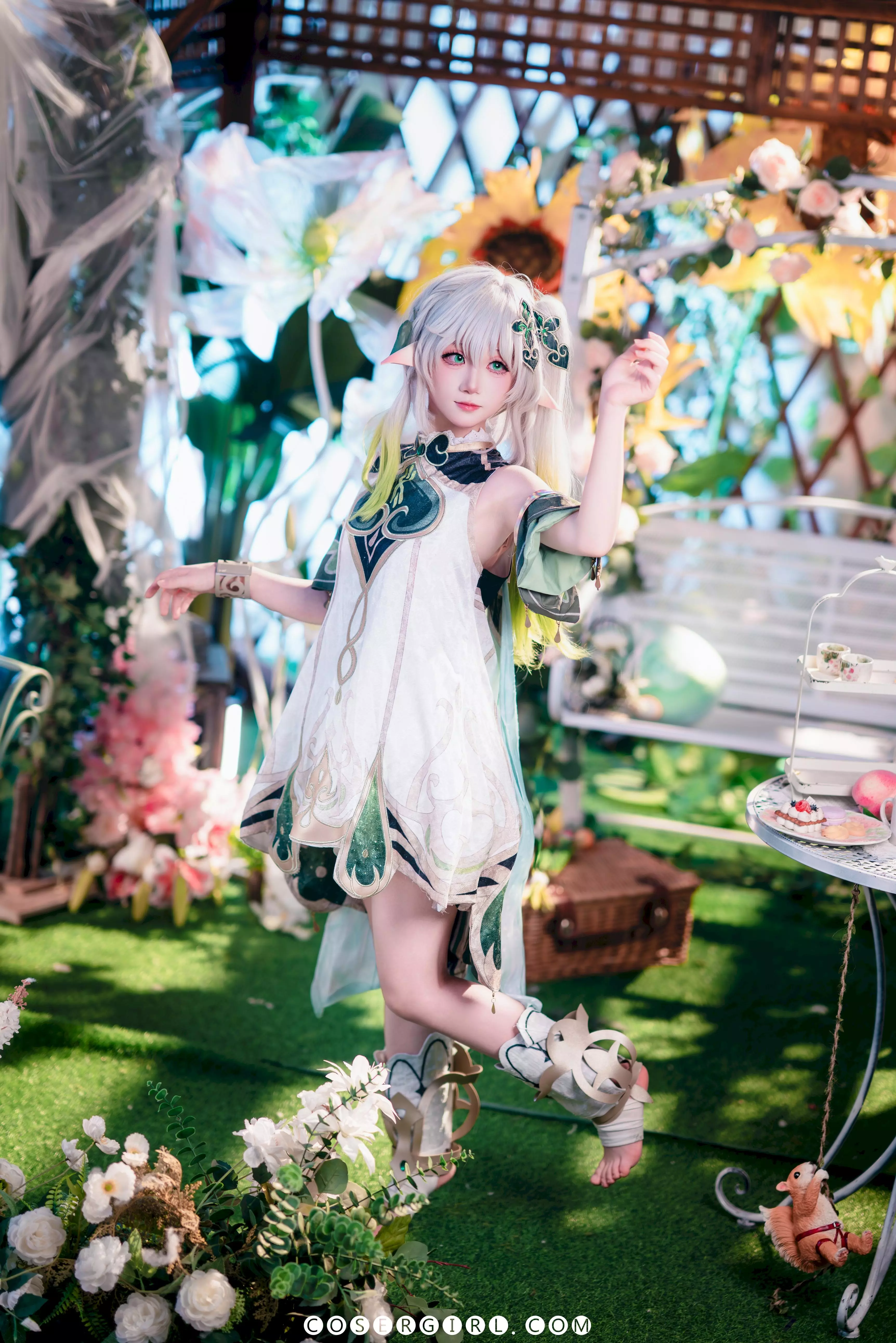 ran3扮演的纳西妲站在花园布景中,手持蓝色飘带,对着镜头展现出甜美的微笑。